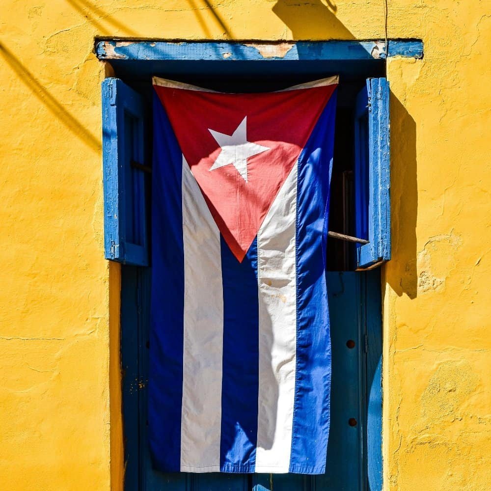 Bandera de Cuba en una ventana con persiana azul y pared amarilla, símbolo de identidad y cultura cubana, en un entorno vibrante y colorido que refleja el espíritu del país.