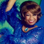 Amac artista en vestuario azul con plumas y joyería llamativa, sonriendo feliz en retrato colorido, promoviendo arte, cultura y diversidad en el ámbito artístico en España.