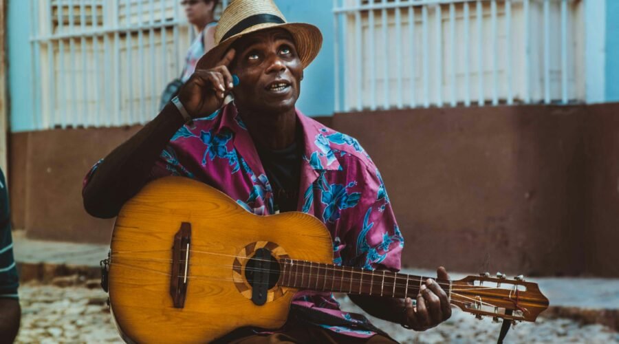 Atractivo artista callejero tocando guitarra, con sombrero y vestimenta colorida, en un entorno urbano en Cuba, promoviendo el arte y la cultura cubana.