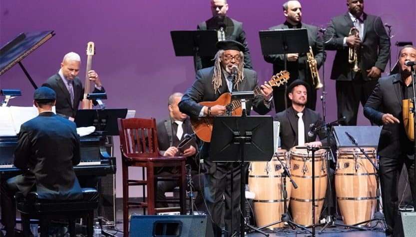 Jazz band en vivo tocando música en un escenario artístico, con instrumentos como guitarra, congas y pianos, en un evento cultural promovido por AMAC en el contexto hispano.
